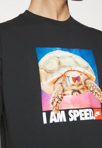 Czarny t-shirt z dużą grafiką żółwia w okularach przeciwsłonecznych na tle galaktyki, z napisem „I AM SPEED.” poniżej.