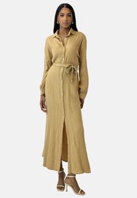 Lang skjortekjole med knapper i lyst beige tekstureret stof, med bindebånd i taljen og sideslids, stylet med hvide sandaler med høje hæle.