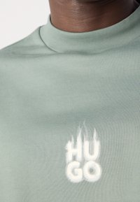 Gros plan d'une personne portant une chemise vert clair avec un logo "HUGO" lumineux sur la poitrine.