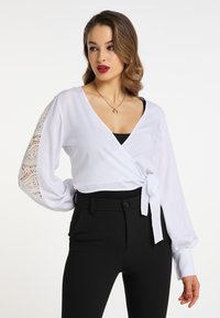 Weißes Wickelbluse mit langen, gepufften Ärmeln und Spitzendetails. Mit gebundenem Taillengürtel, kombiniert mit schwarzen, eng anliegenden Hosen. Glatter Stoff, elegantes Design.