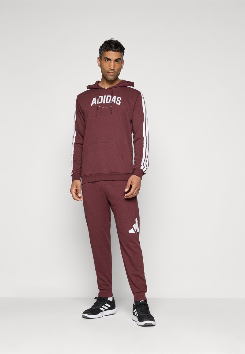 Homme debout portant un hoodie bordeaux Adidas et un pantalon de survêtement assorti avec des baskets Adidas noires et blanches, devant un fond uni.