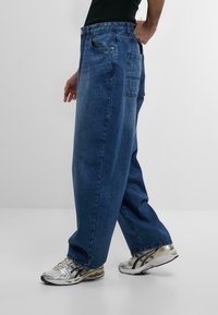 Jeans in denim blu scuro con una vestibilità ampia, dotati di una tasca posteriore e di una leggera scoloritura. Abbinati a scarpe da ginnastica argentate e nere.