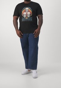Camiseta negra con un gráfico de tres calaveras con acentos coloridos. Llevada con jeans rectos azules y zapatillas blancas.