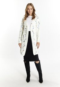 Cardigan bianco e peloso con decorazioni circolari colorate, abbinato a una gonna nera lavorata a maglia e stivali neri alti fino al ginocchio.