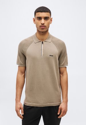 Egy férfi bézs színű, rövid ujjú, elöl cipzáros BOSS logós galléros pólóban, egyszínű háttér előtt áll.