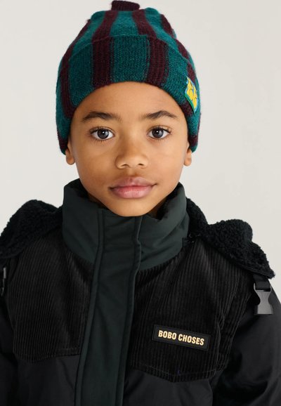 Jeune enfant portant un bonnet tricoté rayé vert et bordeaux et une veste noire avec une étiquette "Bobo Choses" sur la poitrine.