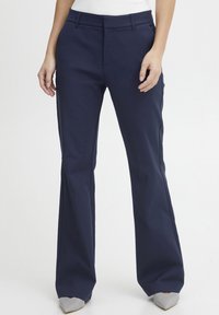 Navy blue flared trousers made of smooth fabric, featuring a flat front, side pockets, and a tailored fit, paired with pointed gray shoes.