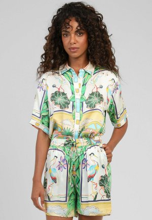 Femme aux cheveux bouclés porte une chemise boutonnée assortie et un short à cordon avec un imprimé coloré d'oiseaux tropicaux et de feuilles.