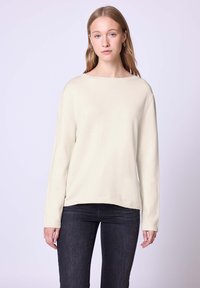 Helles Cremefarbendes Sweatshirt mit einem Bootsausschnitt, langen Ärmeln, gerippten Bündchen und einer lockeren Passform, getragen über dunklen Jeans. Glatte Textur, minimalistisches Design.
