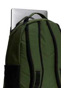 Mochila verde con correas negras parcialmente abierta, una computadora portátil plateada se está colocando dentro del compartimento principal contra un fondo blanco.