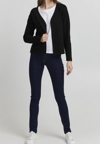 Černý kardigan s dlouhými rukávy a otevřeným předním dílem, nošený přes bílé tričko, v kombinaci s tmavě modrými skinny džínami a bílými teniskami.