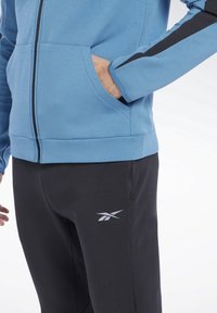 Blå zip-up hoodie med svarta accenter, som har sidofickor. Svarta byxor med en logotyp, som betonar mjukt, slätt tyg och en figurnära design.