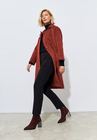 Cappotto in lana color ruggine con colletto su misura e due bottoni, abbinato a pantaloni neri a tapered e stivaletti borgogna con tacco geometrico.