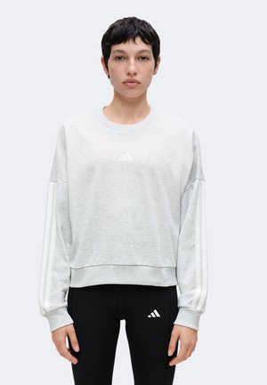 Lühikeste tumedate juustega inimene, kes kannab heledat halli Adidas dressipluusi ja musti Adidas logodega retuuse, seisab otse vastu valget tausta.