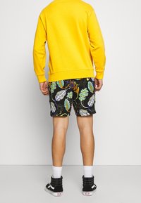 Sweatshirt jaune, shorts noirs avec des motifs de feuilles vifs en vert et violet, et baskets montantes noires. Chaussettes blanches visibles.