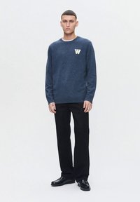 Homme debout, portant un pull bleu marine avec un "W" blanc sur la poitrine, un pantalon noir et des mocassins noirs, sur un fond blanc uni.