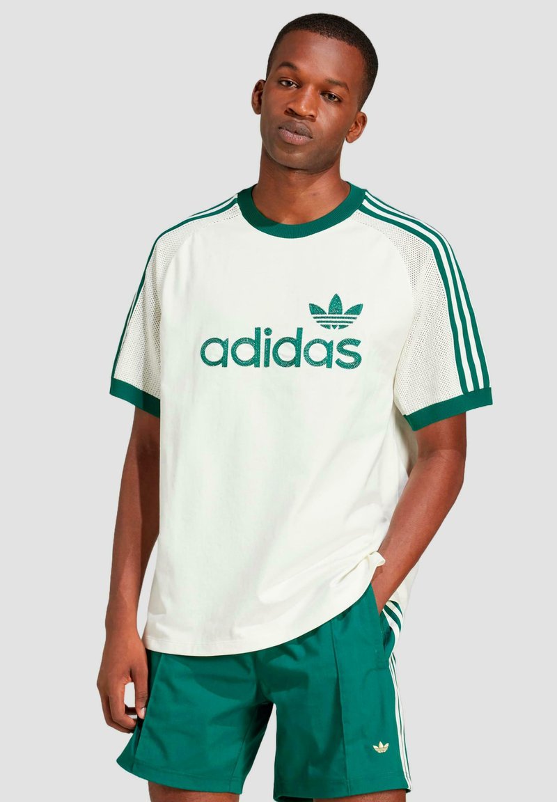 Balta trumpa rankovėmis marškinėliai su žaliais akcentais, turintys tinklelio intarpus, siuvinėtu "adidas" logotipu, ir žali šortai su logotipu ant krašto.