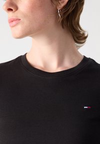 T-shirt en coton noir avec un col rond, arborant un petit logo bleu, blanc et rouge sur la poitrine. Texture lisse, coupe décontractée.