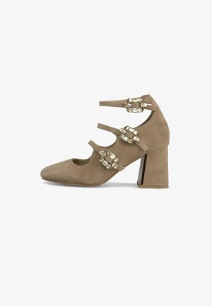 Beige suède schoen met een spitse neus, drie decoratieve banden met kristalaccenten, en een brede blokhak met een metallic detail.