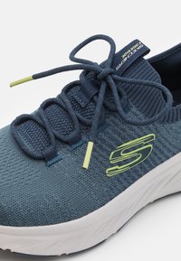 Zapatilla deportiva azul marino con parte superior de malla, ojales reforzados y suela acolchada blanca. Presenta detalles en verde lima y cordones elásticos.