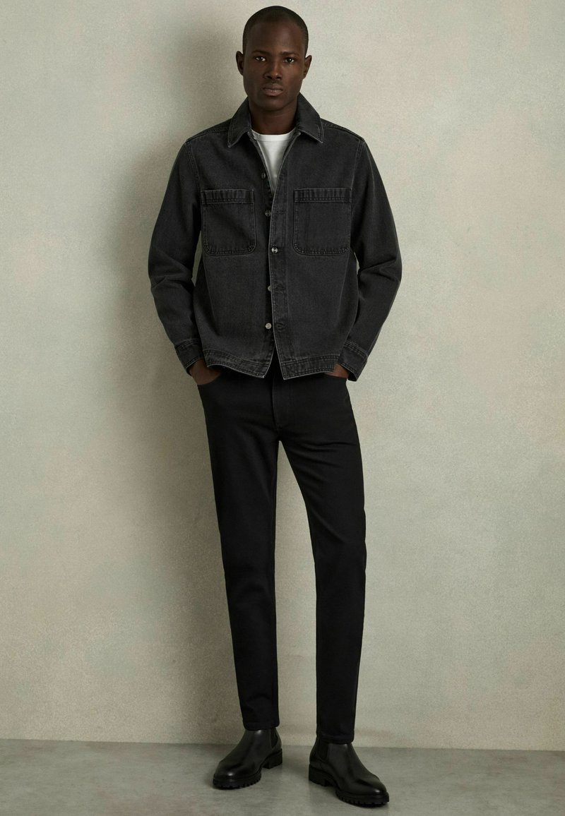 Jachetă din denim gri cu două buzunare la piept, purtată peste un tricou alb, combinată cu pantaloni negri și bocanci negri până la gleznă. Textură netedă, design lejer.
