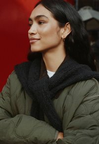 Quilted olivgrüner Mantel mit einem schwarzen Strickschal. Das Model hat lange dunkle Haare und minimale Accessoires und steht vor einem roten Hintergrund.