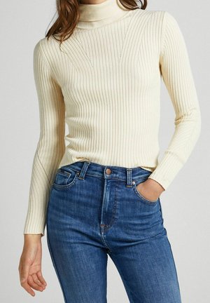 Pull à col roulé côtelé crème avec manches longues, présentant une coupe ajustée, des détails texturés subtils et un col rond, associé à un jean bleu foncé.