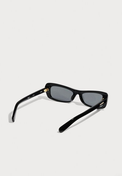 Lunettes de soleil rectangulaires noires avec des verres foncés et des accents dorés sur les branches noires, vues de dos sur une surface gris clair.
