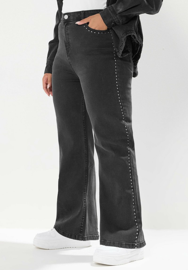 Jeans noirs taille haute avec des clous argentés sur les côtés, portés avec des baskets blanches et une veste noire, main dans la poche.