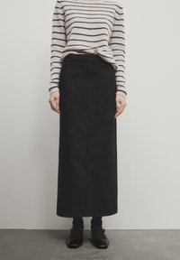 Jupe maxi en denim noir avec deux poches avant, associée à un pull en tricot rayé. La tenue comprend des chaussures sombres et des chaussettes montantes.