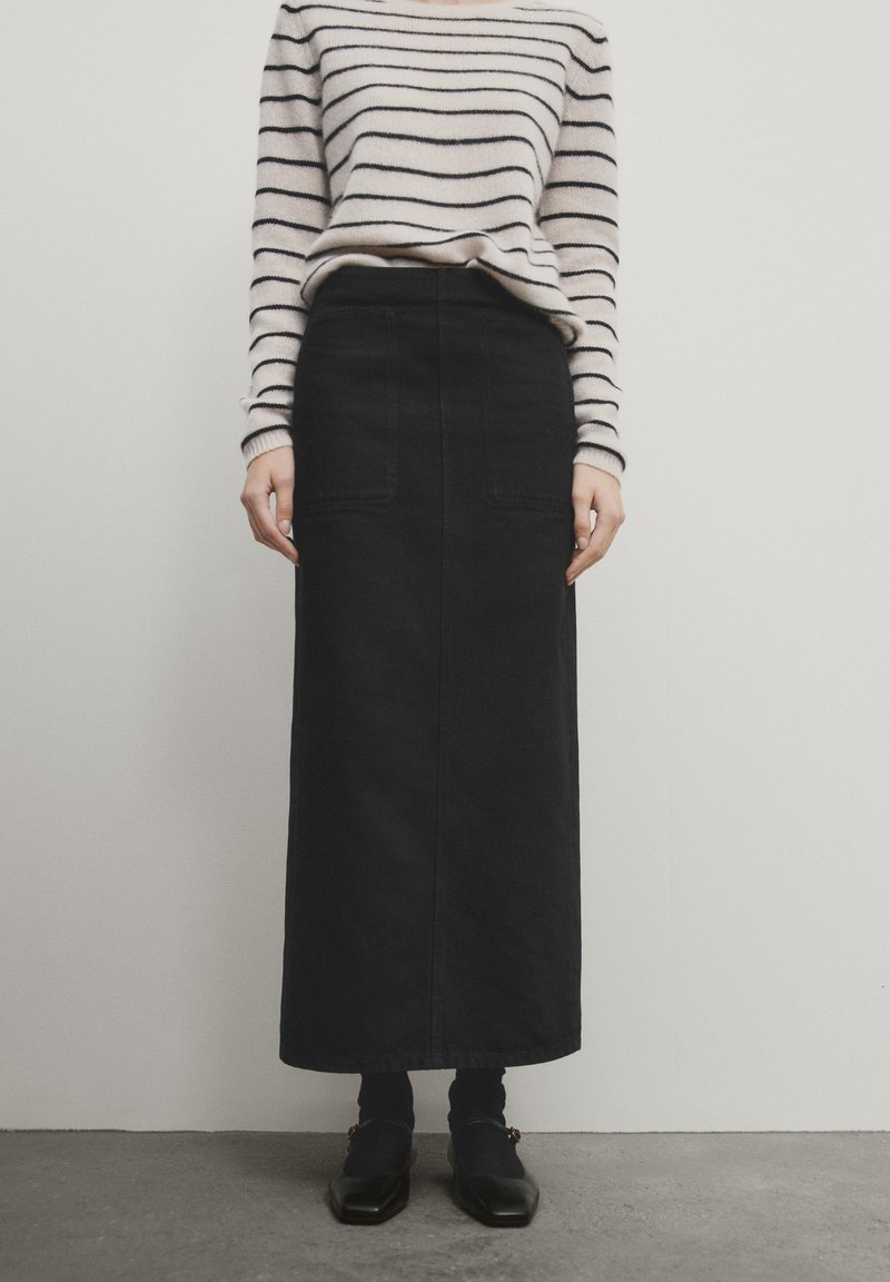 Jupe maxi en denim noir avec deux poches avant, associée à un pull en tricot rayé. La tenue comprend des chaussures sombres et des chaussettes montantes.
