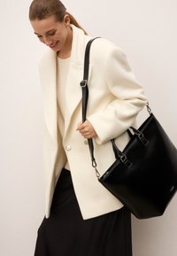 Biely kabát z vlnenej zmesi s veľkými gombíkmi, v kombinácii s krémovým sveterom a čiernou sukňou, drží čiernu koženú tašku s kovovými detailami.