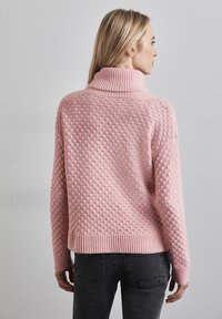 Rosa stickad tröja med hög ribbad krage, strukturerat mönster och ribbade ärmmuddar. Bakom har den en avslappnad passform och diskret detaljer.