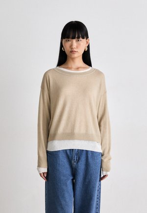 Une jeune femme aux cheveux noirs raides porte des boucles d'oreilles en or, un pull beige sur une chemise à manches longues blanche et un jean bleu ample.