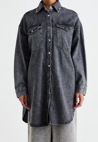 Abito camicia in denim nero a maniche lunghe con bottoni a pressione frontali e tasche a falda sul petto, indossato sopra una gonna grigia.