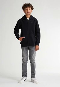 Sort hættejakke med lynlås og frontlomme, båret med lysegrå jeans og hvide sneakers. Har hætte og minimal branding på brystet.