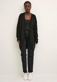 Zwarte gebreide cardigan met geribbelde manchetten en een open voorzijde, gedragen over een zwart shirt en gecombineerd met donkere spijkerbroek en witte sneakers.