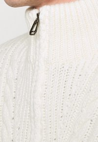 Stickad vit tröja med hög krage, med en halv dragkedja och ett strukturerat flätstickat mönster.