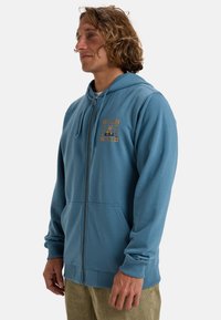 Blue zip-up hoodie made of soft fabric, featuring a kangaroo pocket, ribbed cuffs, and a graphic print on the left chest.