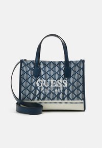 Granatowa i biała torba tote z plecionki z teksturowanym wzorem, skórzanymi akcentami, podwójnymi uchwytami górnymi oraz odpinanym paskiem na ramię. Zawiera logo "GUESS".