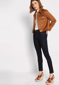 Bruine suède cropped jas met drukknoopsluiting, donkere denim skinny jeans en tan kleurige wedge sandalen met kruislings bandjes.