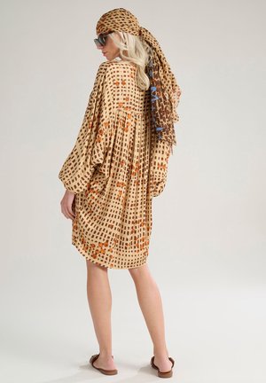 Robe légère beige à motif grille brun et orange, avec une coupe ample, des manches longues et des détails plissés. Associée à un foulard à motifs.