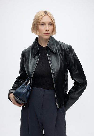 Mujer rubia con corte de pelo bob llevando una chaqueta de cuero negra, top negro, pantalones azul marino y sosteniendo un pequeño bolso azul oscuro.