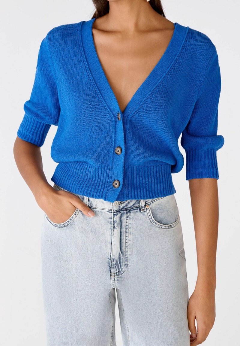 Femme portant un cardigan boutonné bleu vif à manches trois-quarts et un jean taille haute délavé clair, main dans la poche.