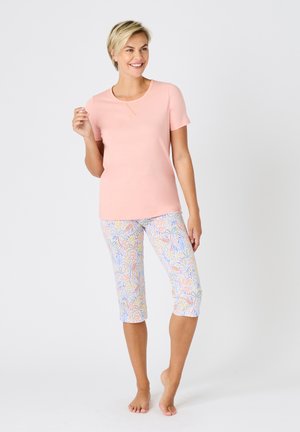 Mujer sonriente de pie descalza, con una camiseta de manga corta rosa claro y pantalones capri blancos con un patrón floral colorido.