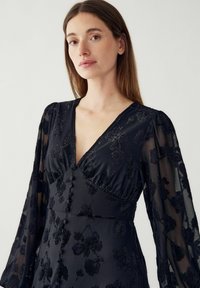 Czarna sukienka w kwiaty z przezroczystymi długimi rękawami, głębokim dekoltem w serek, detalami z guzikami oraz teksturowanym materiałem z haftowanymi wzorami.