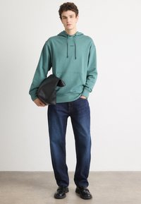 Felpa con cappuccio in teal, con tasca frontale, abbinata a jeans dritti blu scuro e scarpe nere, tenuta con una borsa nera.