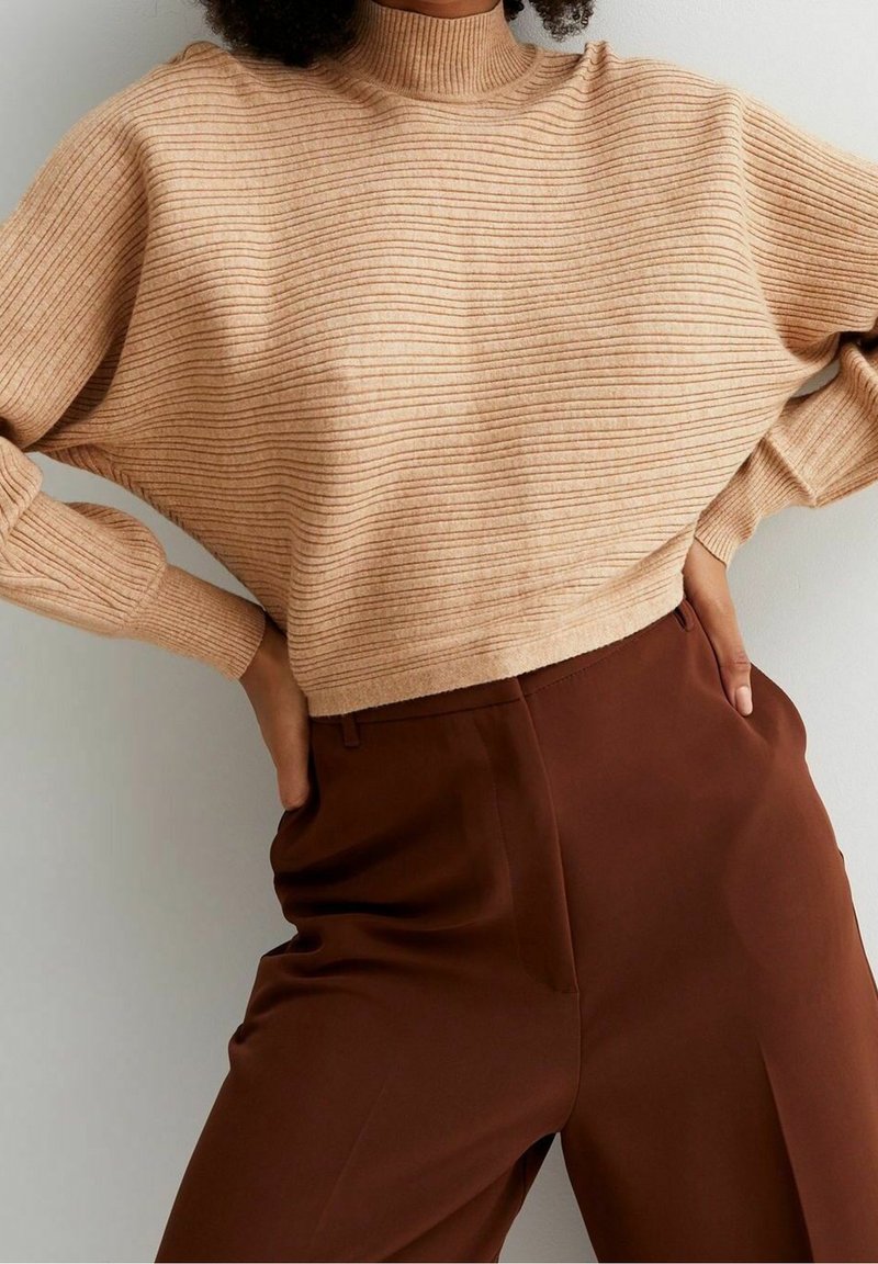 Personne portant un pull à col roulé côtelé beige et un pantalon taille haute marron foncé, les mains posées sur les hanches.