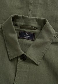 Olivovo zelená košeľa na gombíky s čiernym gombíkom a čiernou štítkom so slovami „Matinique Heritage Apparel“ prišitým vo vnútri.