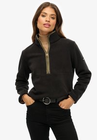 Pull en polaire noir avec col montant et demi-zip, présentant une bordure vert olive et un détail de couture sur la poitrine, associé à un pantalon noir.